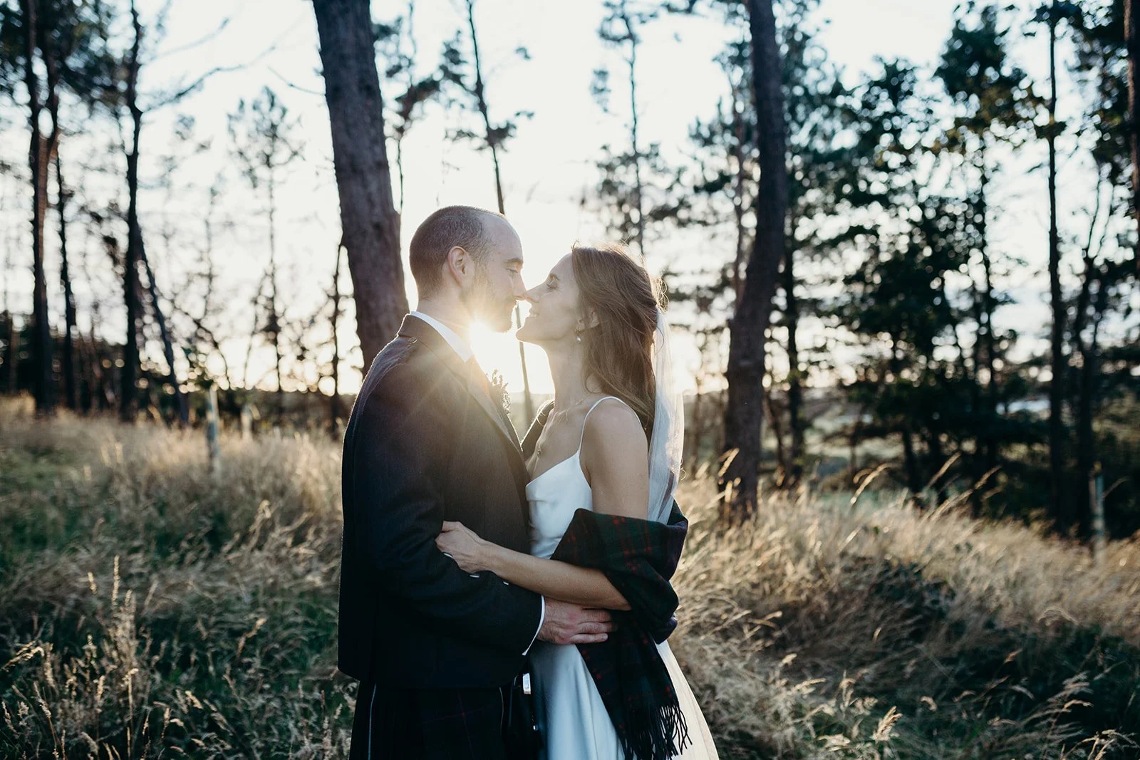 A bride and groom standing close in a forest at sunset, about to kiss, with the sun shining behind them.