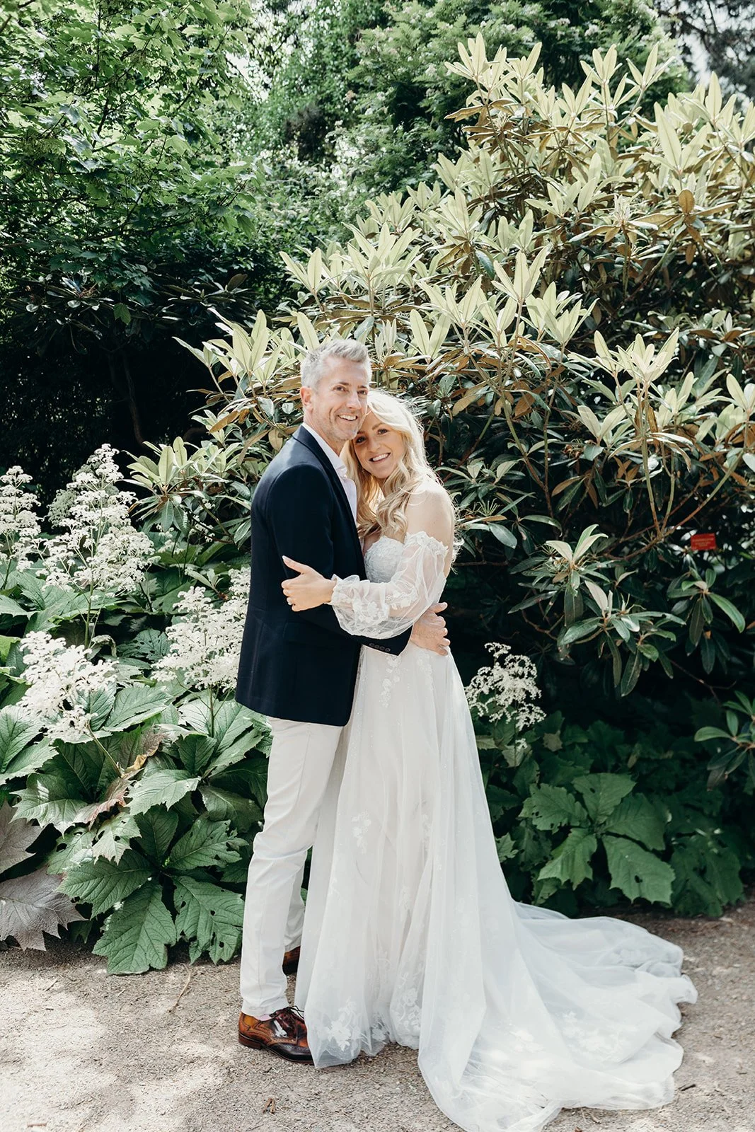 A newlywed couple smiling and embracing outdoors surrounded by green foliage and flowering plants.