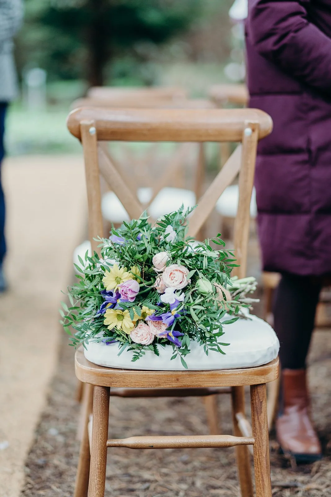 A bouquet of flowers with pink, yellow, purple, and white blossoms, placed on a wooden chair with a white cushion, at an outdoor event.