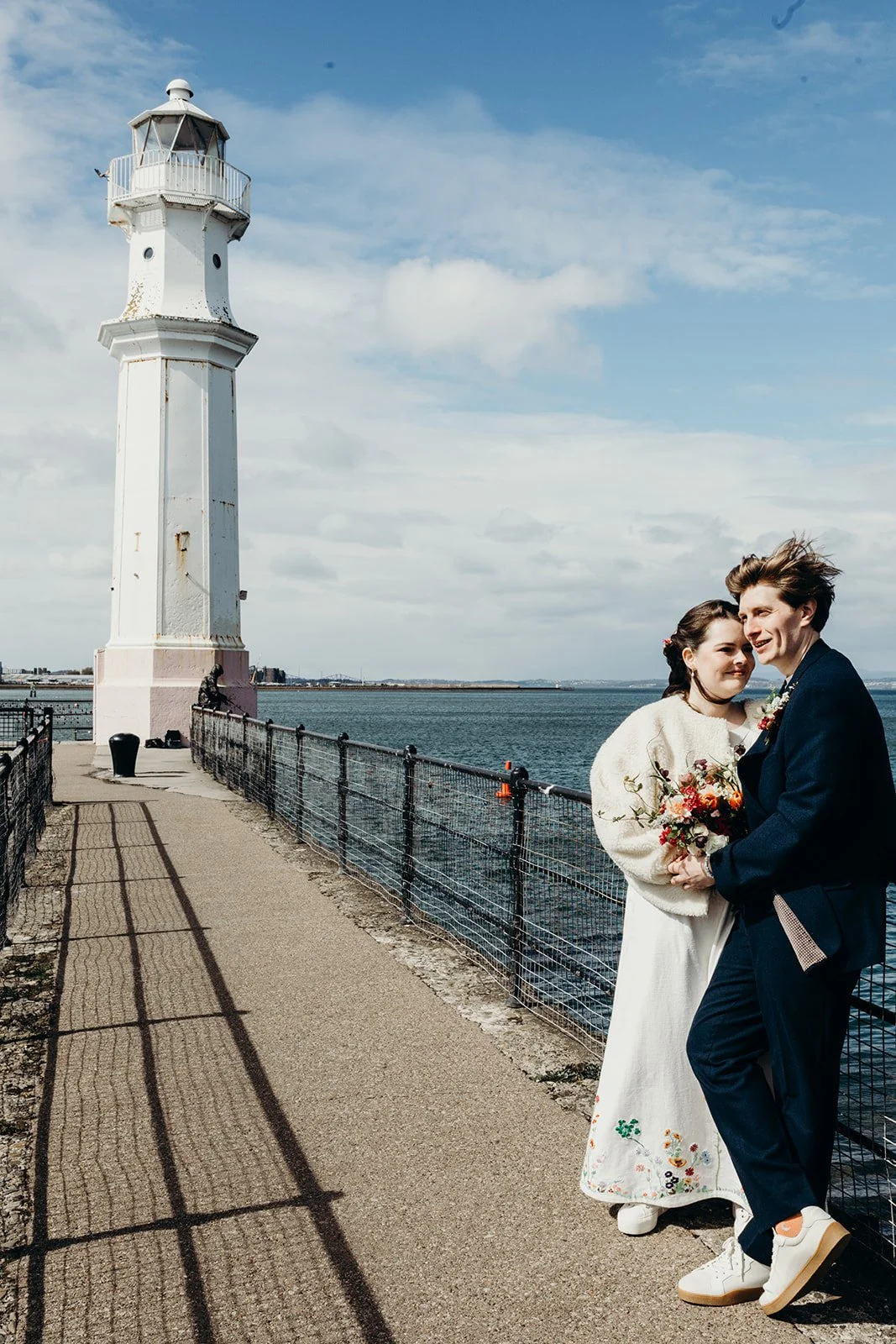 A young couple dressed in wedding attire standing by the waterfront near a white lighthouse on a partly cloudy day.
