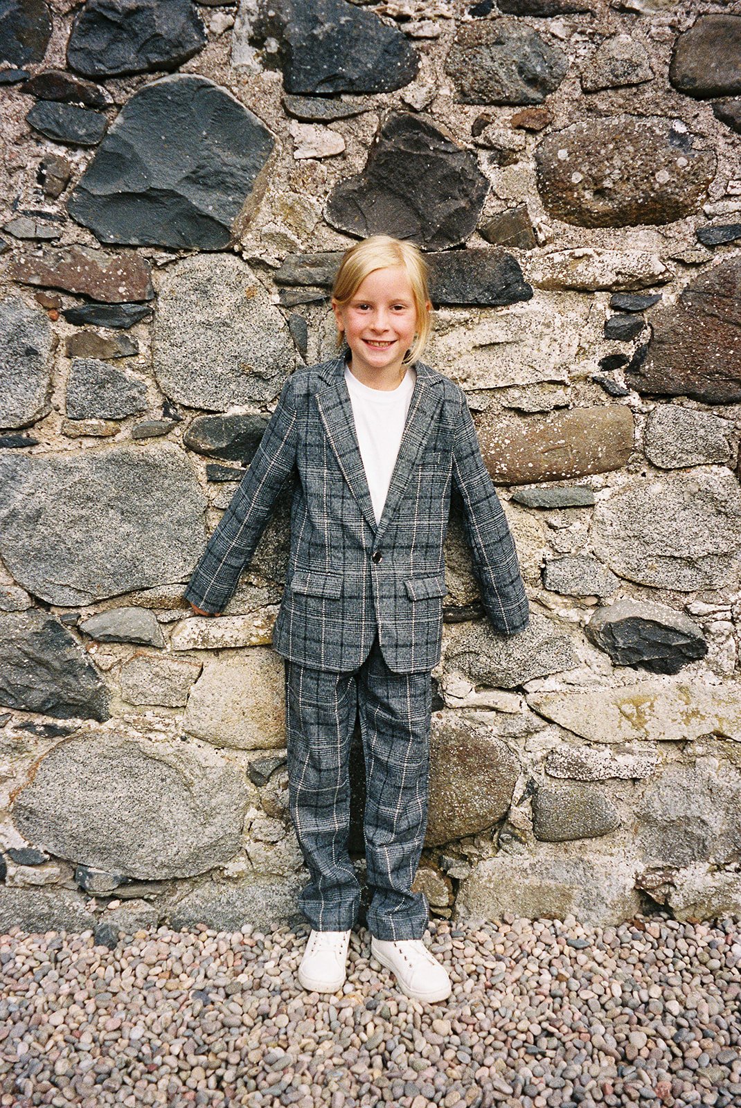A young girl with blonde hair, smiling, standing against a stone wall, wearing a plaid suit and white sneakers.