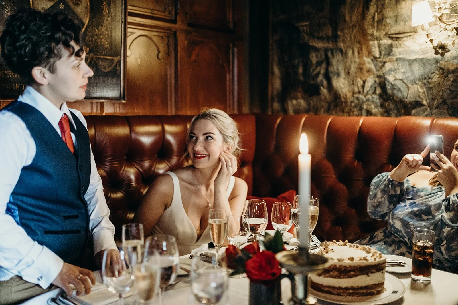 A woman with blonde hair smiling at a server during a dinner in a restaurant, with a woman taking a photo and a cake on the table.