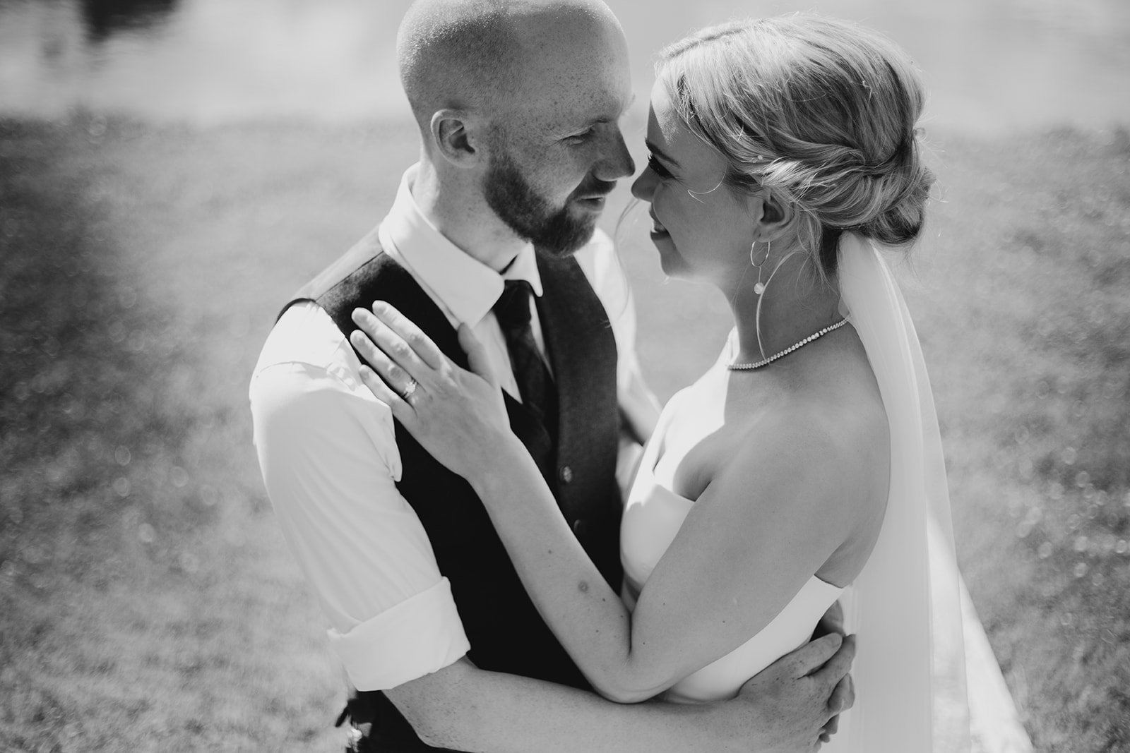 A black and white photo of a newlywed couple smiling and holding each other outdoors, with the groom wearing a vest and tie, and the bride in a wedding dress with jewelry.