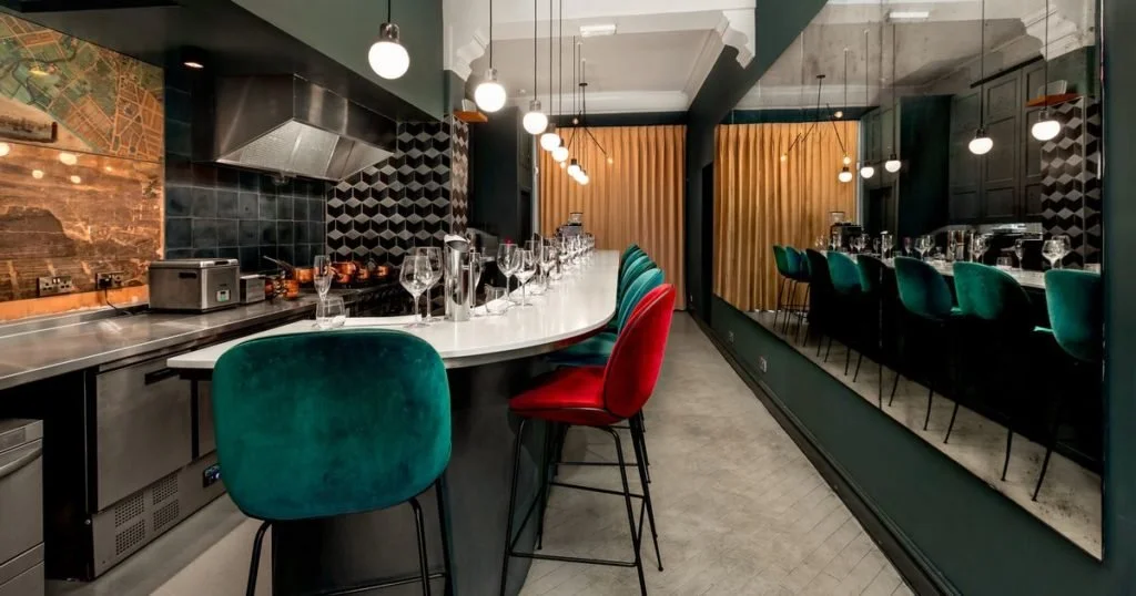 Interior of a modern restaurant or bar with a curved white countertop, colorful chairs, hanging lights, black tiled backsplash, and decorative wall accents.
