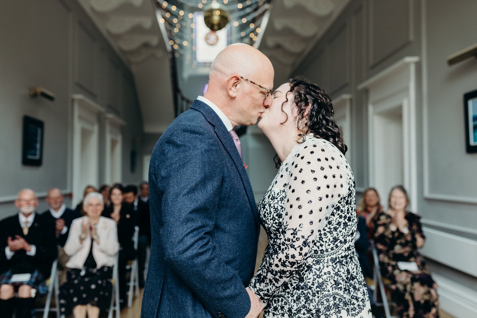A couple kissing at a wedding ceremony, with the bride and groom holding hands and guests watching in a decorated indoor venue.
