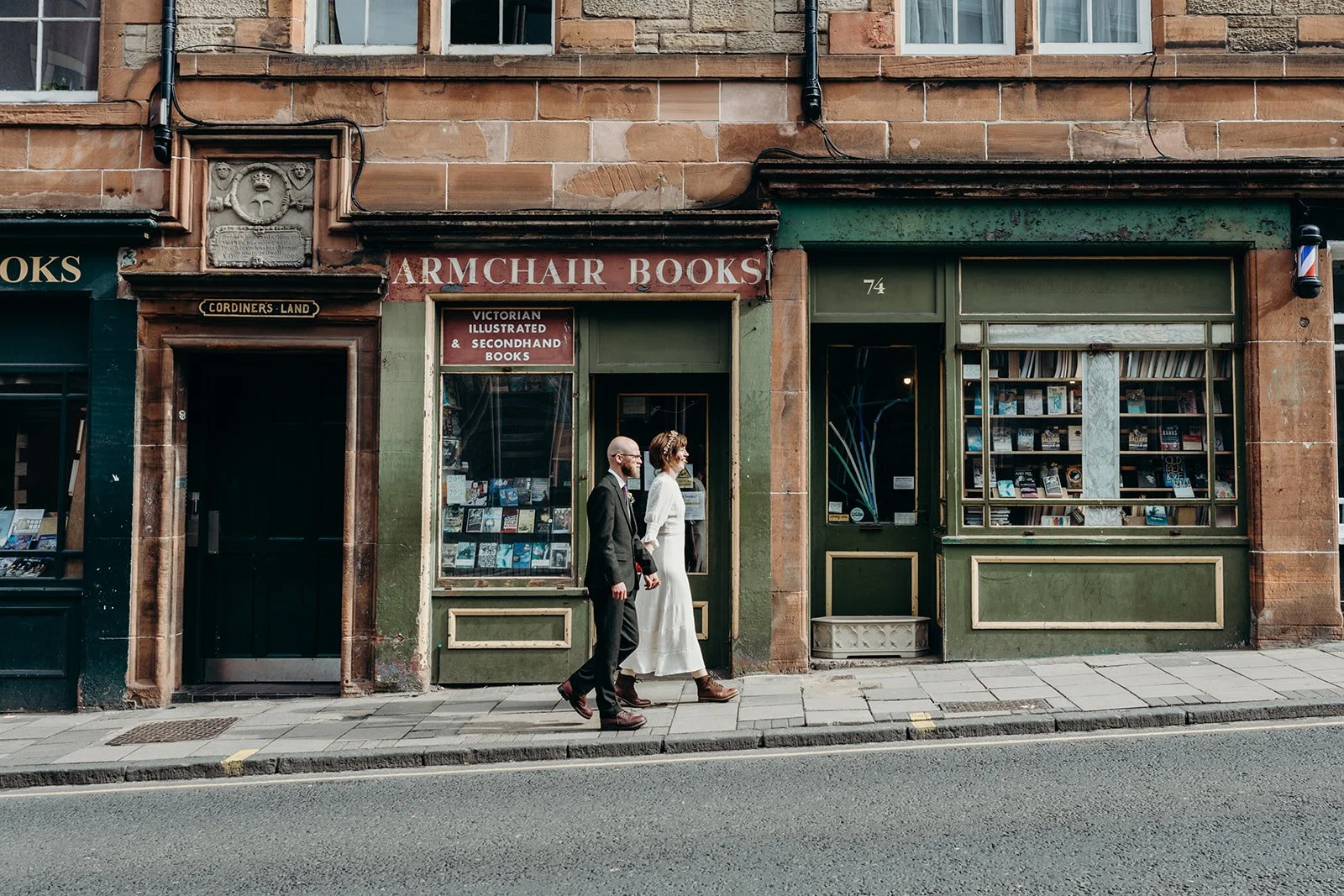 Two people walking past a vintage bookstore with a green facade and large display windows filled with books, on a city sidewalk.