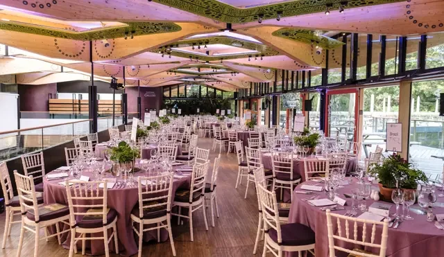 Wedding reception banquet hall with round tables covered in mauve tablecloths, decorated with potted plants, glassware, and cutlery, set for guests, with large windows and modern ceiling design.