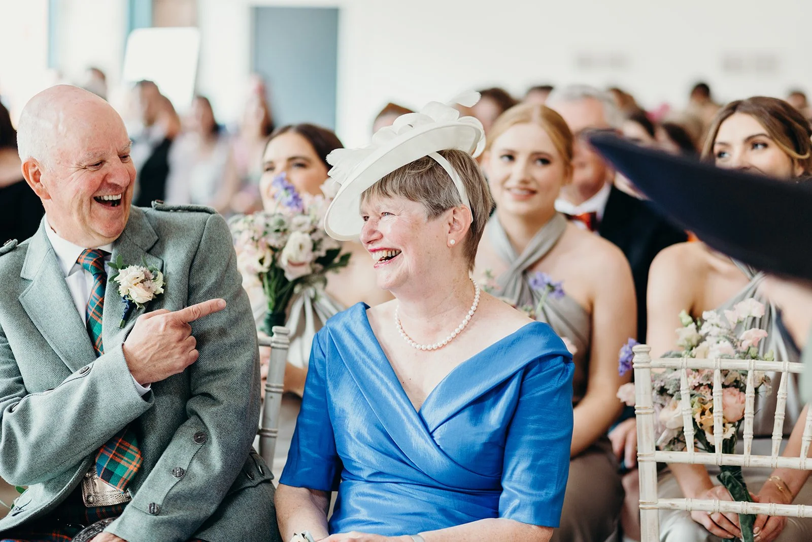 Smile of an elderly woman in a blue dress and white hat, sitting next to a man in a gray suit at a wedding.
They are surrounded by bridesmaids holding bouquets, all smiling and talking.