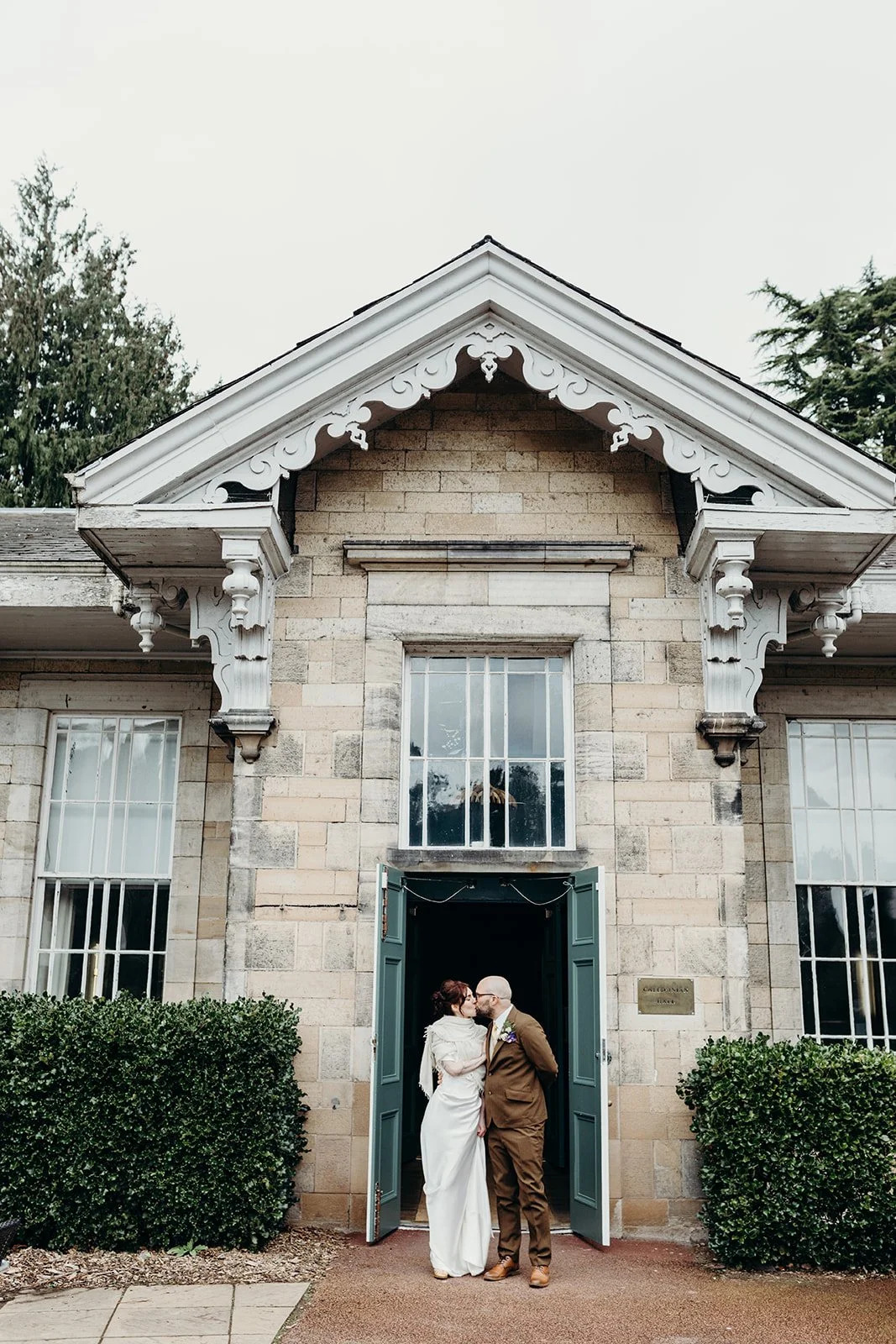 A couple dressed in wedding attire sharing a kiss in front of a historic stone building with open doors, surrounded by green bushes.