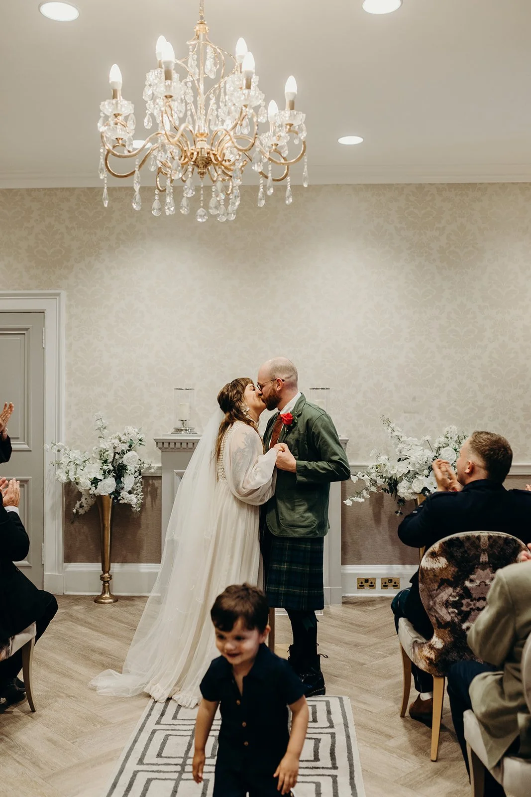 A bride and groom share a kiss during their wedding ceremony in a decorated indoor venue with guests watching and clapping.