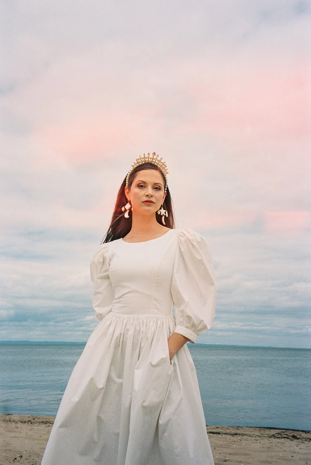A woman in a white dress standing on a beach at sunset, wearing a golden crown and star-shaped earrings.