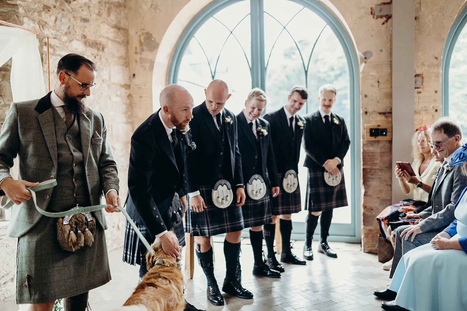 A group of groomsmen dressed in matching Scottish kilts and jackets, bowing during a wedding ceremony inside a rustic stone building with large arched windows, while guests seated nearby observe.