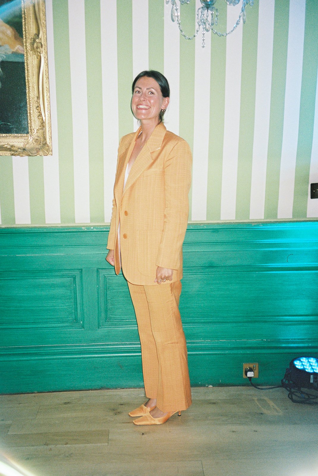 Woman in a yellow suit standing indoors in front of striped green and white wallpaper, with a painting and chandelier overhead.