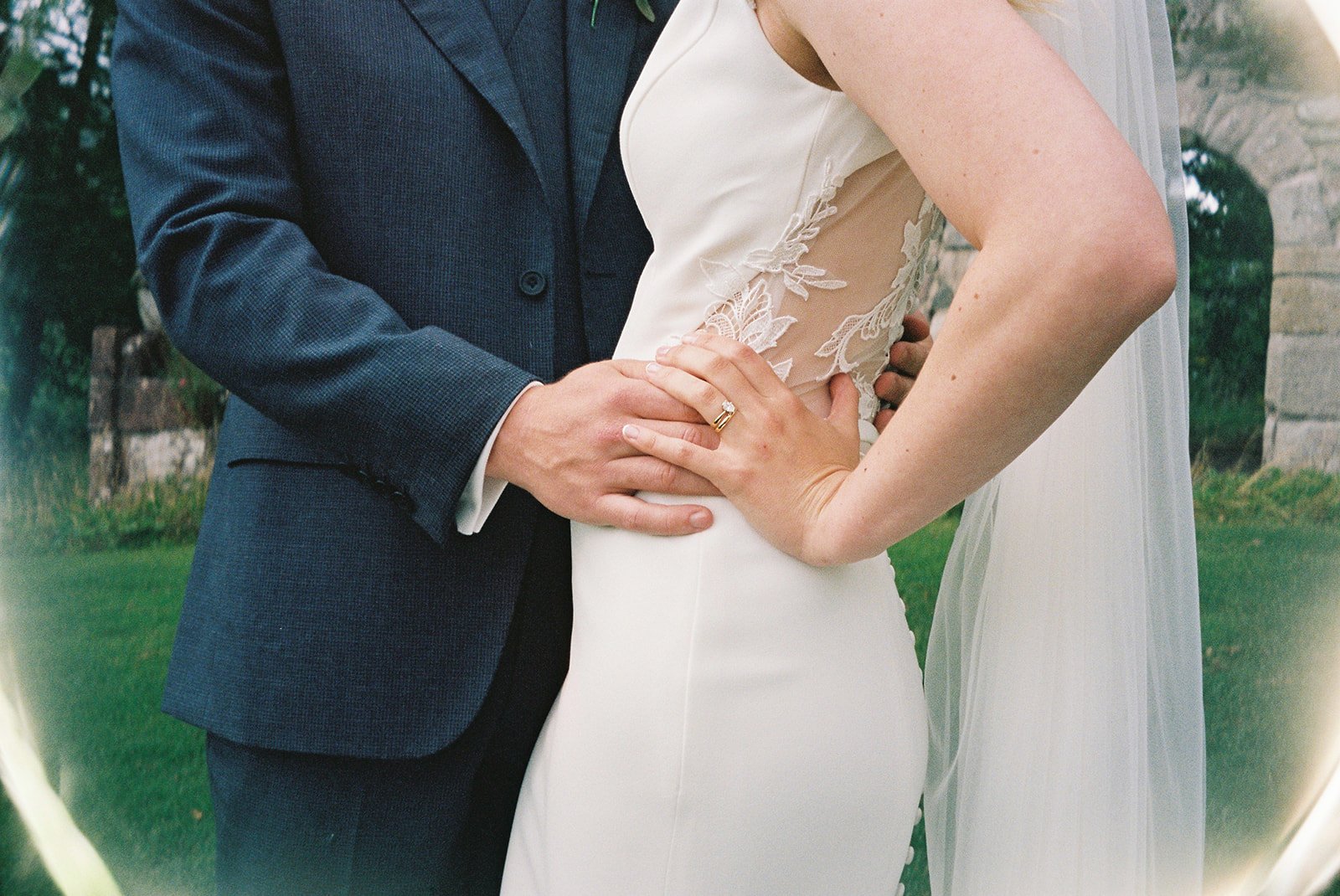 Close-up of a bride and groom at their wedding, showing the couple holding their hands with wedding rings, with the bride wearing a white dress with lace details and the groom in a navy suit.