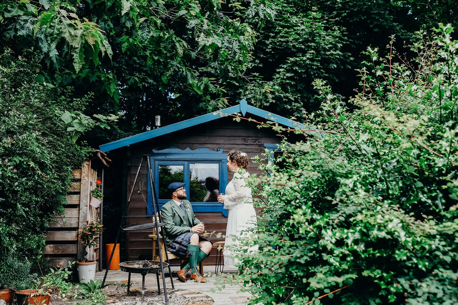 A man sitting on a bench in a garden talking to a woman standing nearby, with a small garden shed and surrounded by lush green foliage.