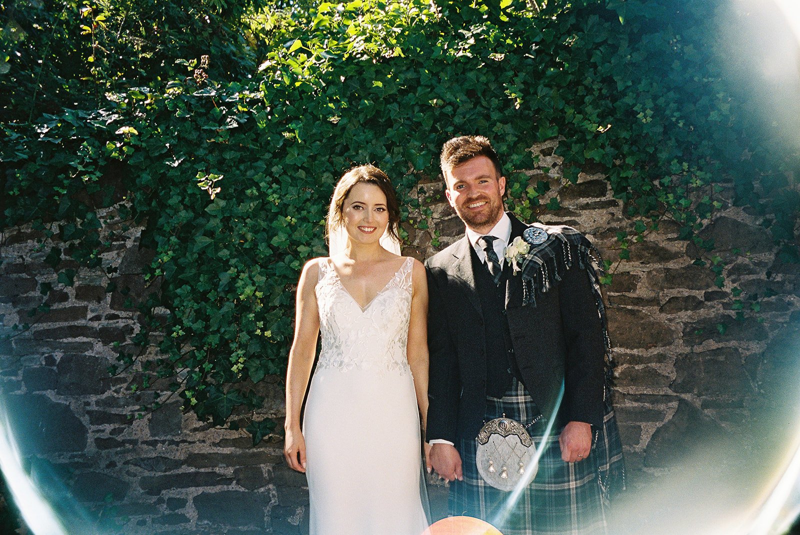A bride and groom standing hand in hand outdoors in front of a stone wall covered with ivy, smiling. The bride is in a white lace wedding dress, and the groom is dressed in traditional Scottish kilt and jacket.