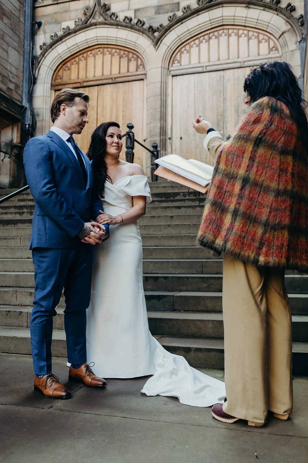 A couple holding hands during a wedding ceremony in front of a historic building, officiant reading from a book.