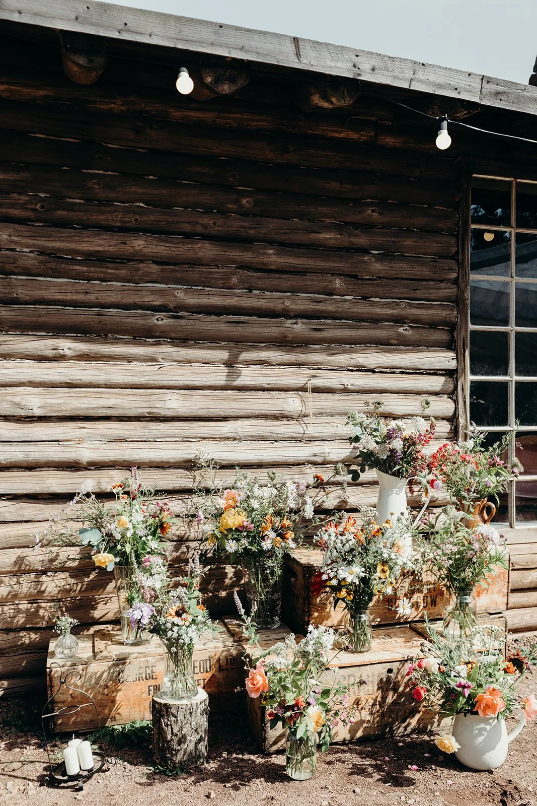 Arrangement of various colorful flowers in vases and jugs placed on wooden crates outside a rustic wooden building with ladder and string lights.