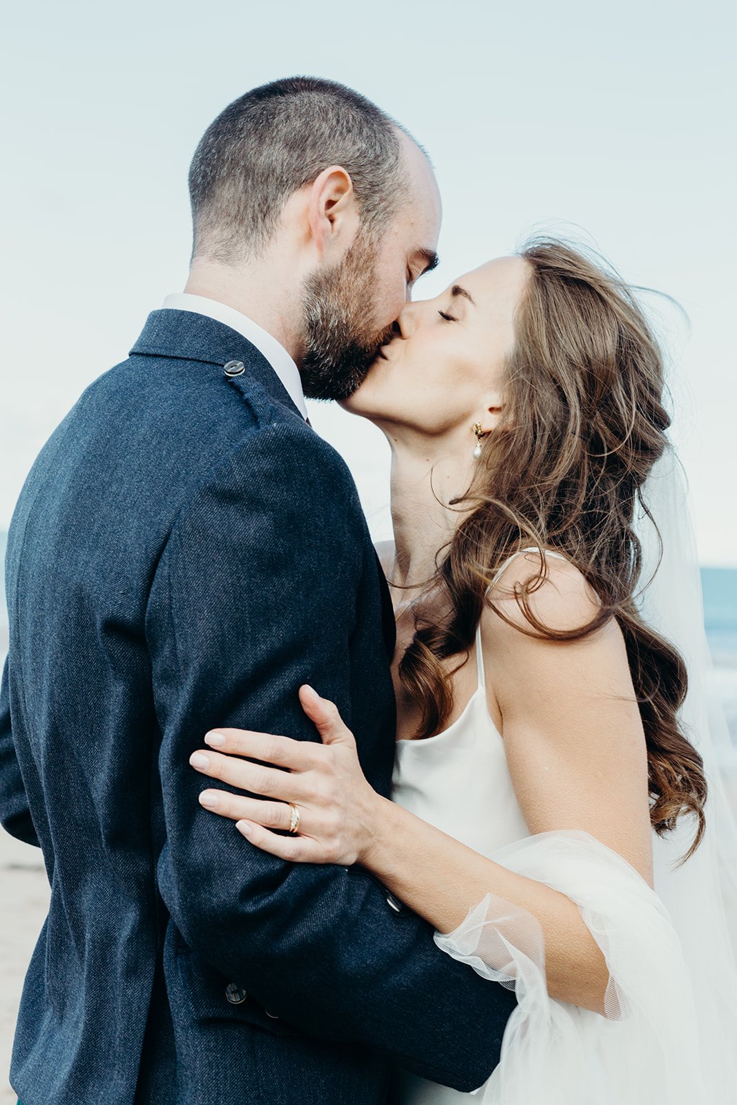 A couple dressed in wedding attire sharing a kiss outdoors, with the ocean in the background.