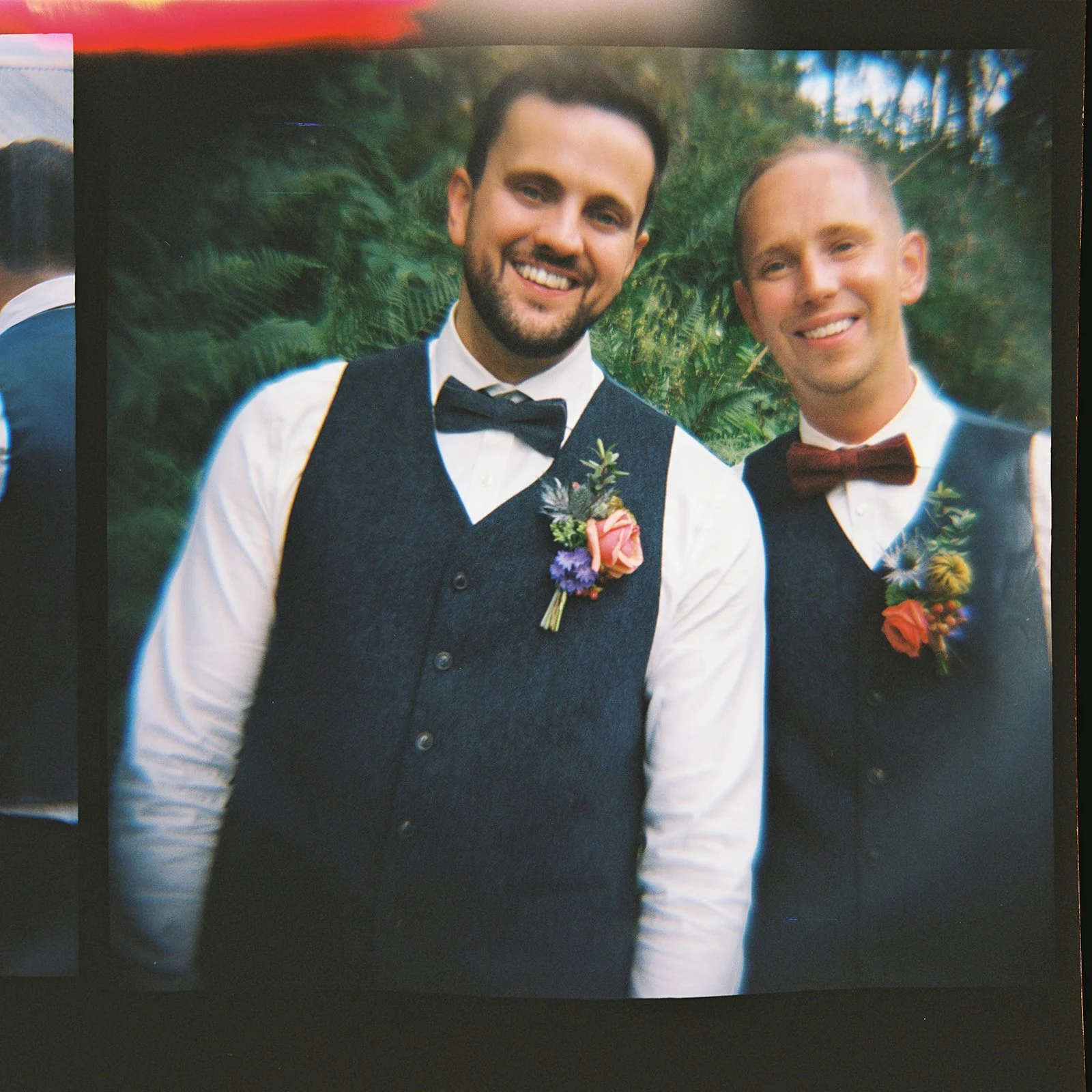 Two men dressed in formal attire with bow ties and boutonnières, smiling outdoors with green foliage in the background.