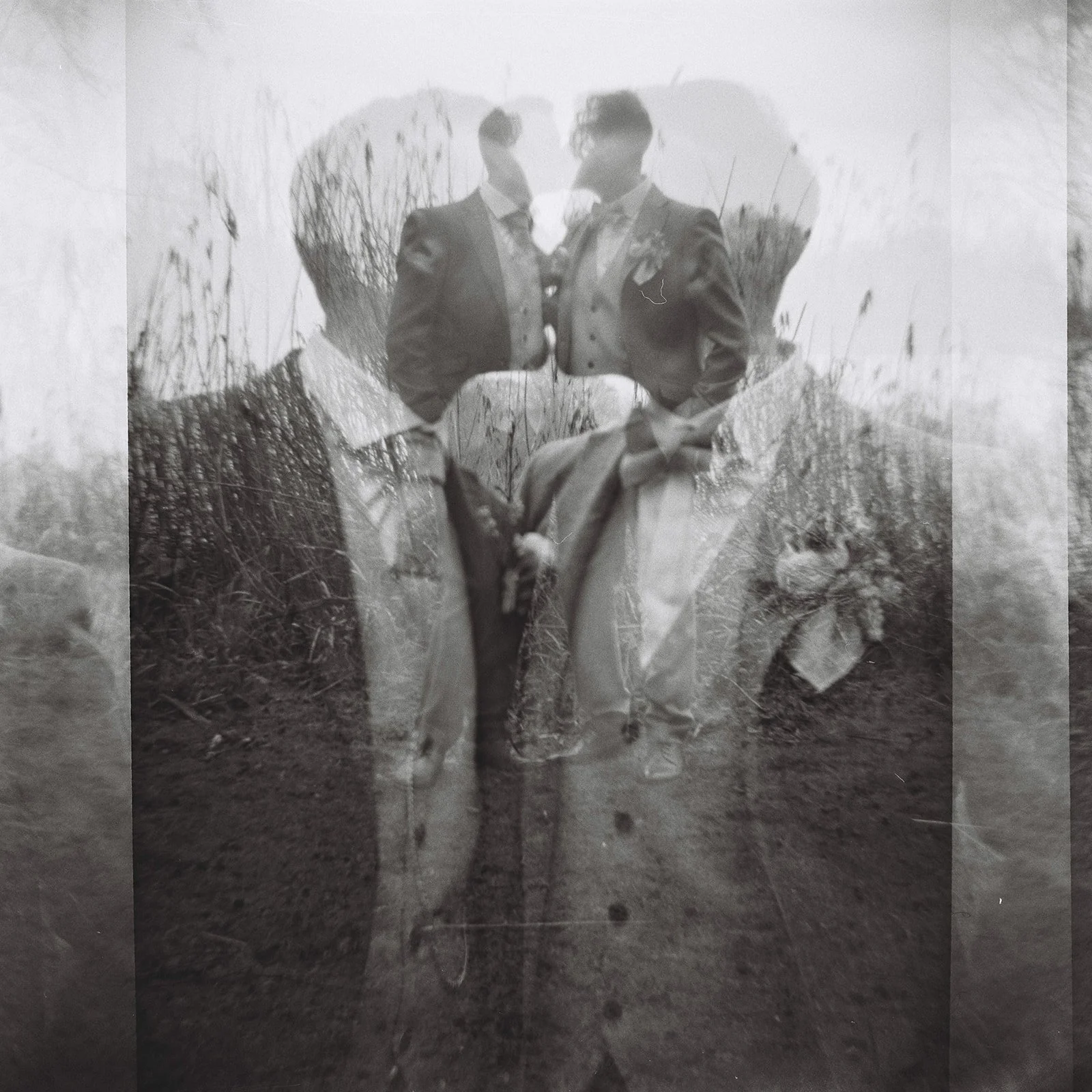 Overlaid black and white photograph of two men in suits standing outdoors with tall grass and plants in the background.