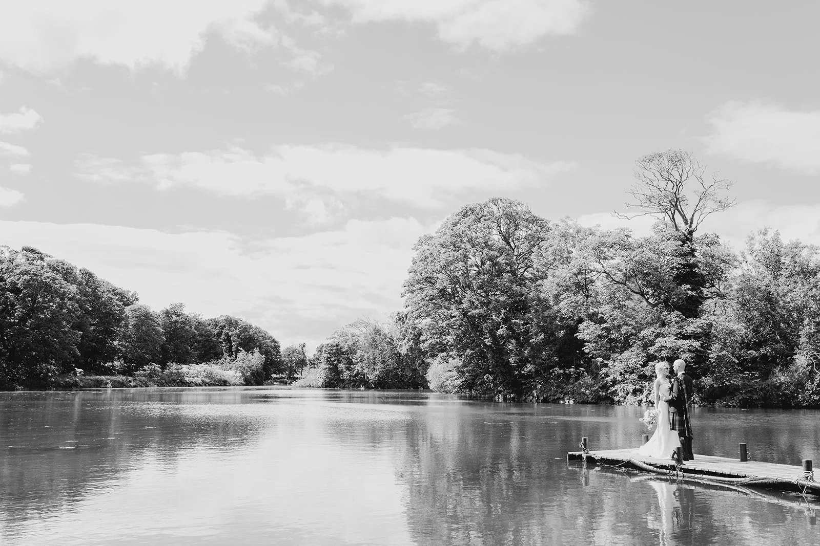 A black and white photo of a couple in wedding attire standing on a small dock by a calm river, surrounded by trees with a partly cloudy sky above.