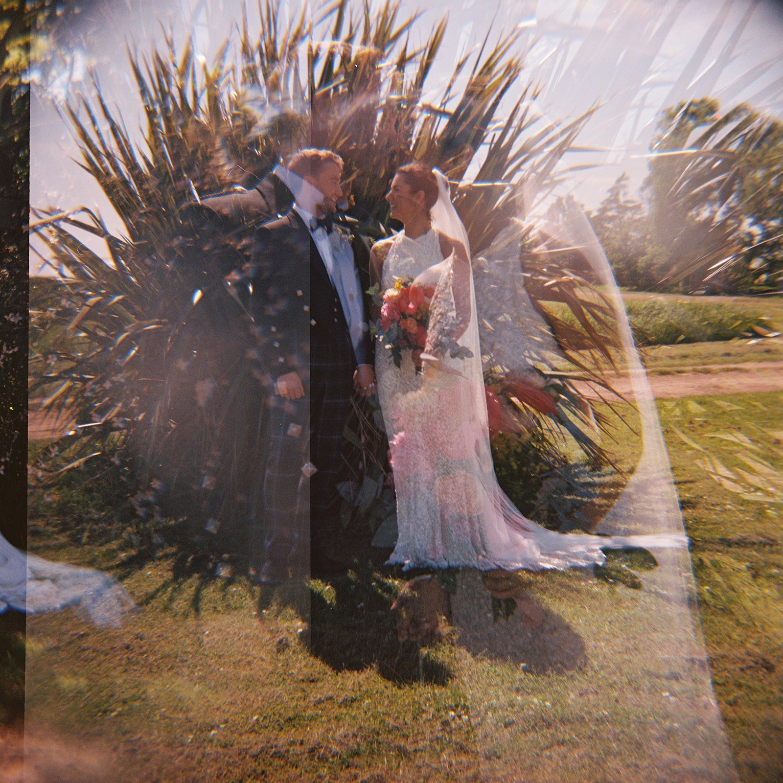 A wedding couple standing outdoors near tall plants, sharing a moment. The bride is in a white lace dress holding a bouquet, and the groom in a black tuxedo with a checkered kilt. The photo has a double exposure effect with a faint image of a glass o