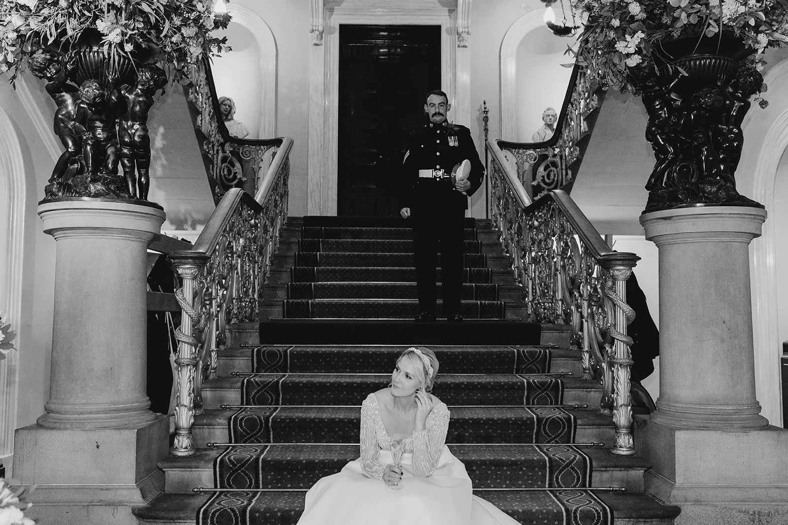 A woman in a white dress with long sleeves sitting on staircase in a formal setting, with a soldier in a decorated uniform standing at the top of the stairs behind her.