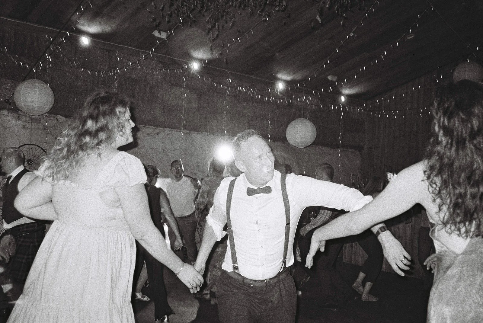 A group of people dancing at a celebration or party in a rustic venue with string lights and paper lanterns hanging from the ceiling.