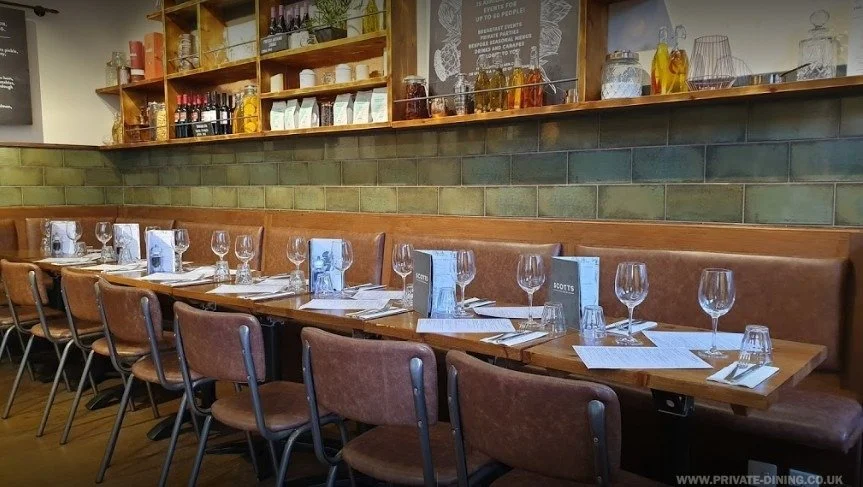A long restaurant table set for dining with about a dozen chairs, glasses, napkins, and menus. The background features a green tiled wall and wooden shelves holding bottles and decorative items.