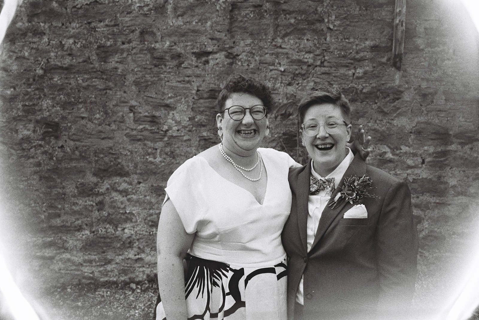 Black-and-white photograph of two women smiling and posing together in front of a stone wall. One woman is dressed in a patterned skirt and blouse with pearl necklaces, and the other is in a suit with a boutonnière, bow tie, and glasses.