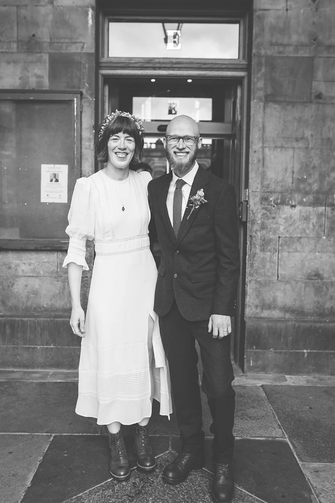 A couple, dressed in wedding attire, standing outside a building, smiling.