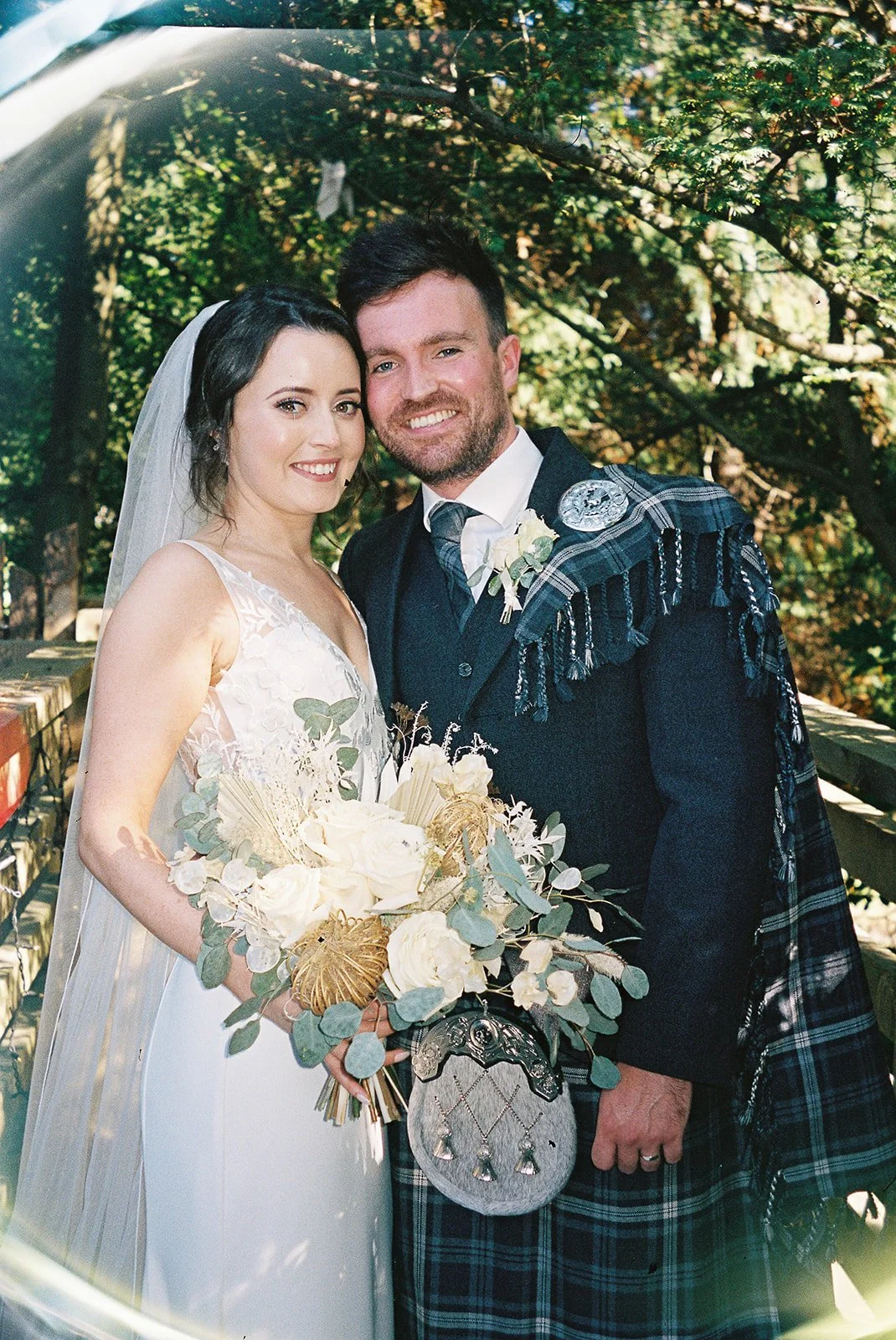 A newlywed couple standing outdoors on a wooden bridge surrounded by trees, smiling at the camera. The bride is wearing a white wedding dress and veil, holding a bouquet of white and green flowers. The groom is dressed in traditional Scottish attire 