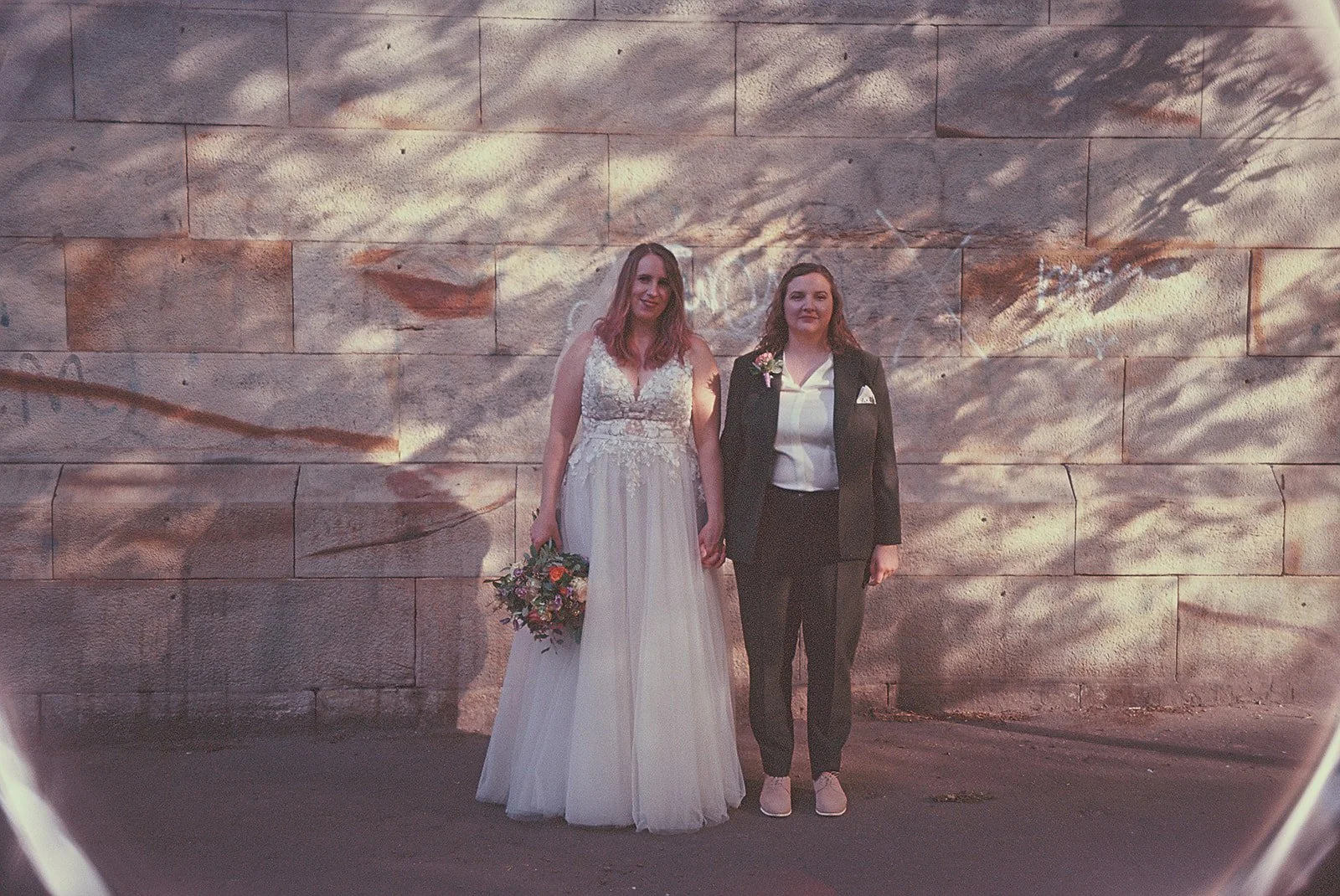 Two women standing side by side, holding hands, in front of a large stone wall. One woman is dressed in a white wedding gown, holding a bouquet, and the other is in a black suit with a white shirt and pink shoes. They are outdoors with sunlight and s