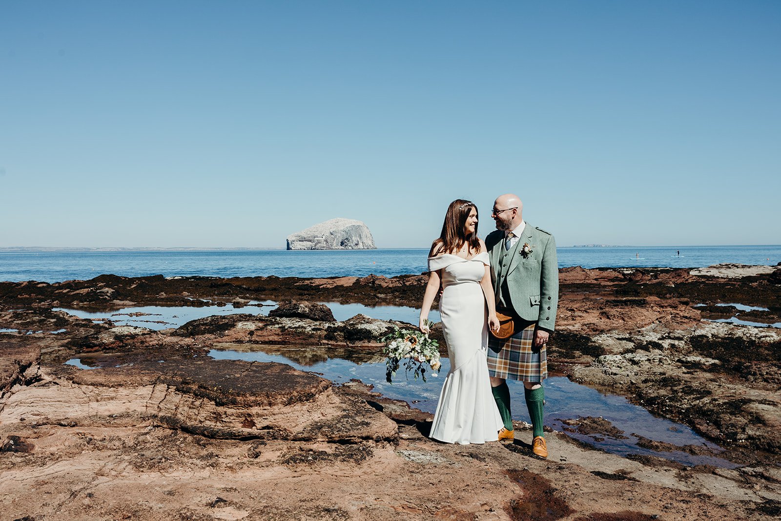 A bride in a white wedding dress holding a bouquet standing next to a groom in a green jacket, plaid kilt, and green socks on a rocky beach with the ocean and a large rock formation in the background on a clear day.