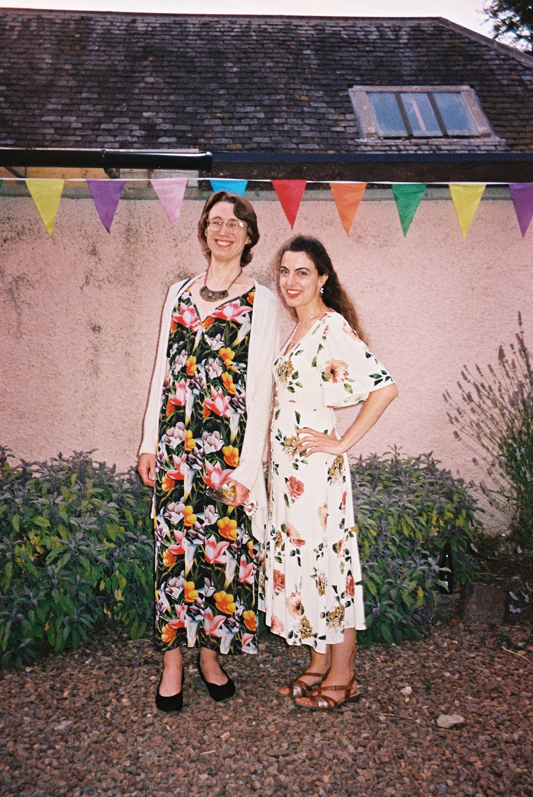 Two women in floral dresses standing side by side outdoors, smiling, with rainbow-colored bunting above them and a house with a shingled roof in the background.