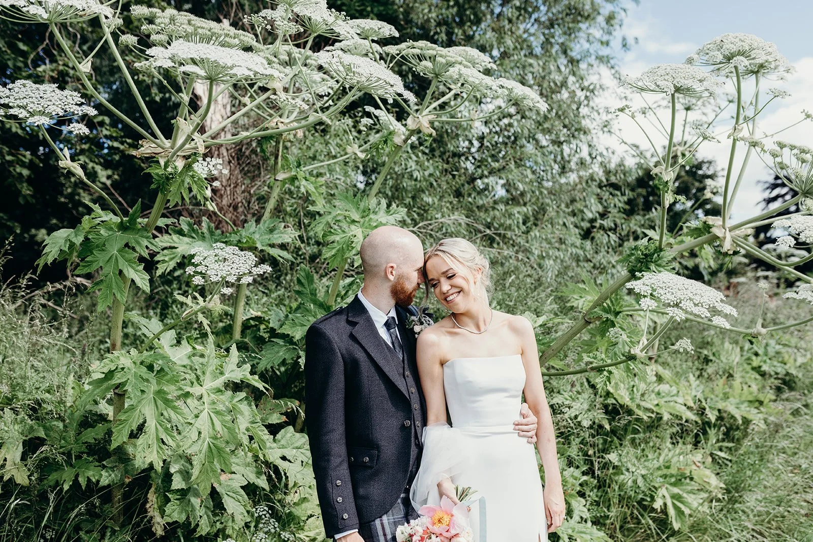 A newlywed couple sharing a joyful moment outdoors, surrounded by lush green plants and large white flowering plants, with a backdrop of trees and partly cloudy sky.