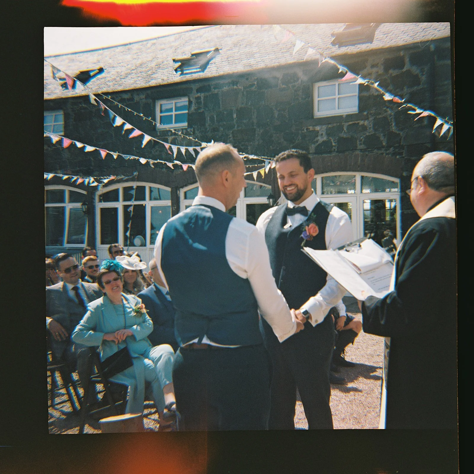 A couple of men in wedding attire holding hands and smiling during their wedding ceremony outdoors, with guests seated in the background and colorful bunting decorations overhead.