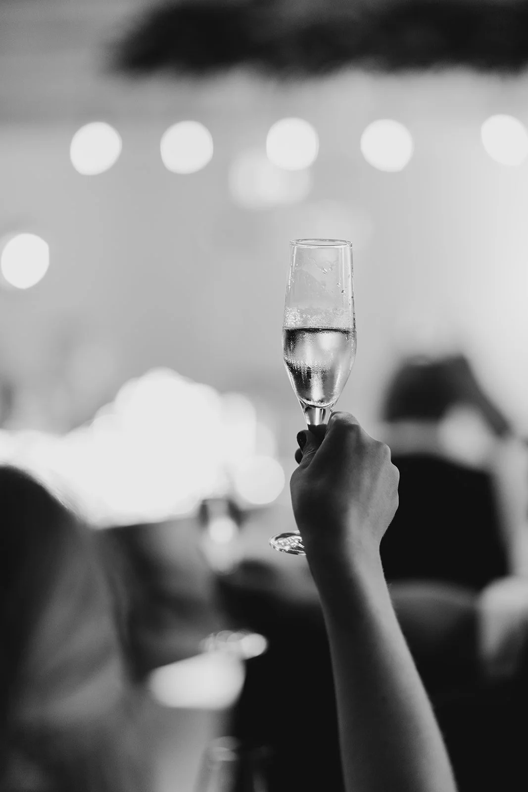 A person holding up a glass of champagne at a celebration or event, with blurred lights in the background.