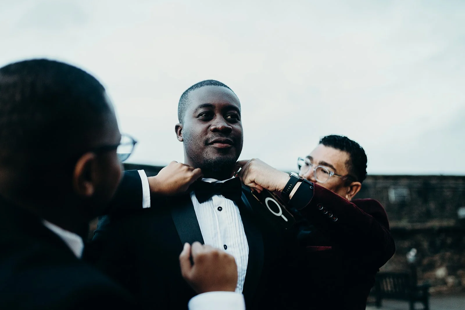 Three men in tuxedos, two adjusting the third man's bow tie outdoors under a cloudy sky.