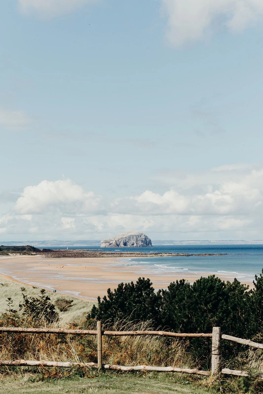A scenic view of a coastal beach landscape with sand dunes, green bushes, and a wooden fence in the foreground. The ocean with gentle waves and a large rock formation or island in the distance under a partly cloudy sky.
