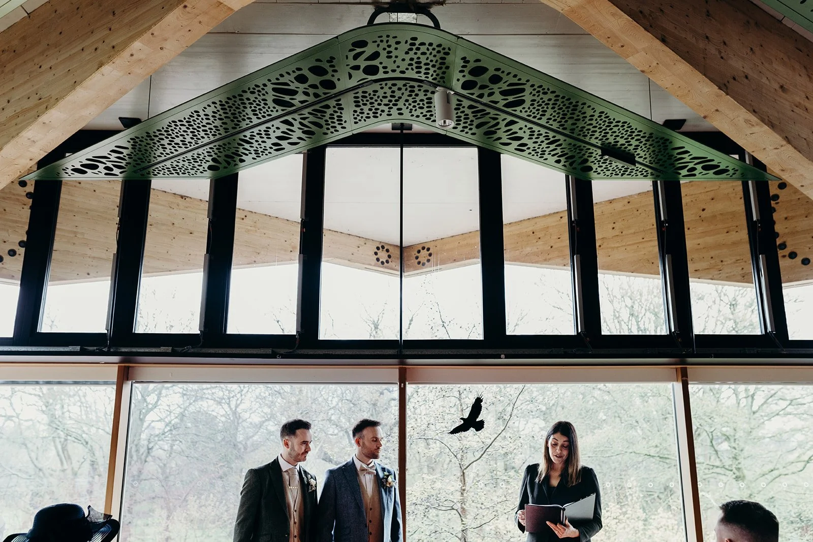 A wedding ceremony taking place indoors with three people standing in front of a large window, and a woman reading from a book or notes. The room has wooden accents and a ceiling with a green perforated panel and a mirrored wall.