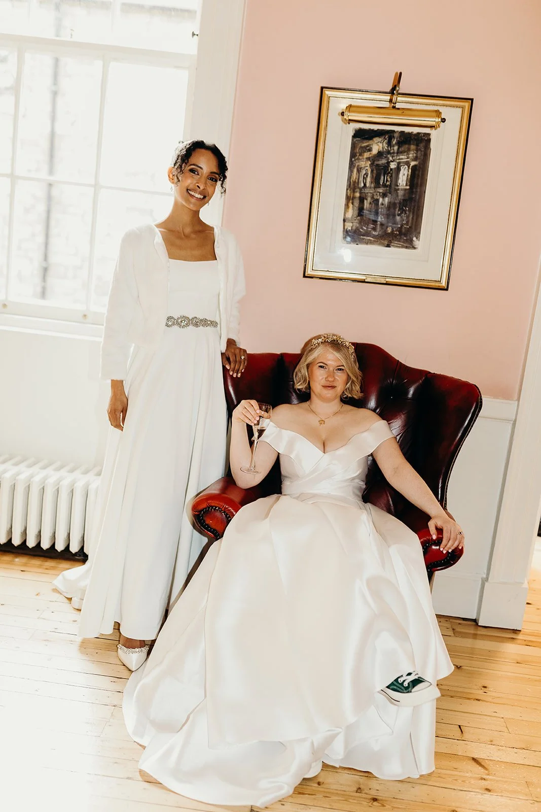 Two women in white dresses at a wedding, one sitting on a red armchair and the other standing beside her, both smiling inside a well-lit room with pink walls and a framed picture above.
