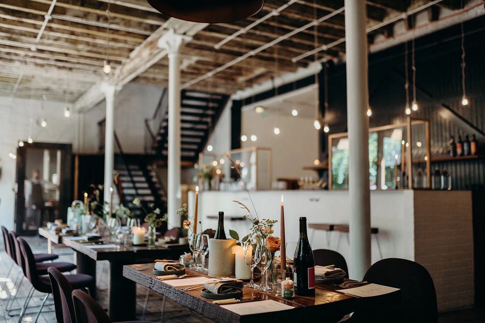 A decorated dining table set for a gathering in a rustic, industrial-style restaurant or event space with candles, flowers, and wine bottles.