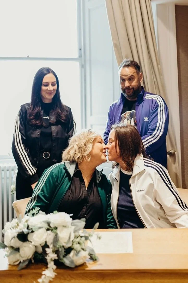 Two women are about to kiss, seated at a table with a bouquet of white flowers. Two other people, a man and woman, stand behind them, observing. All are dressed in athletic jackets.