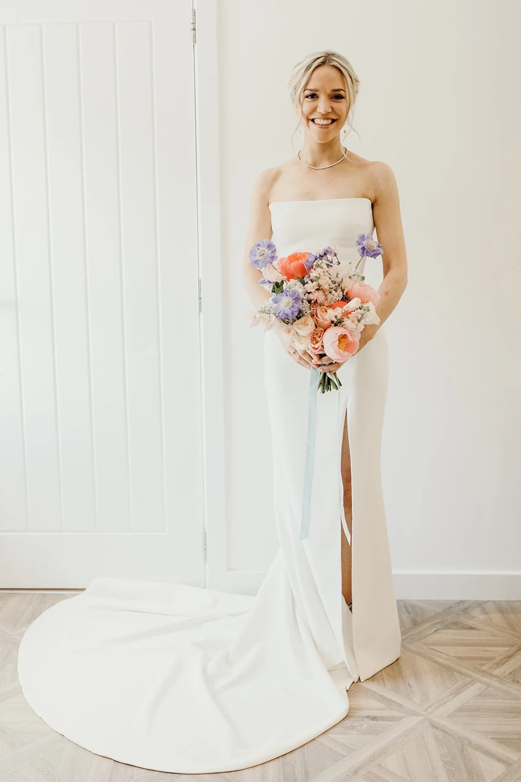 A smiling woman in a white strapless wedding dress holding a colorful bouquet of flowers, standing indoors against a white wall and wooden floor.