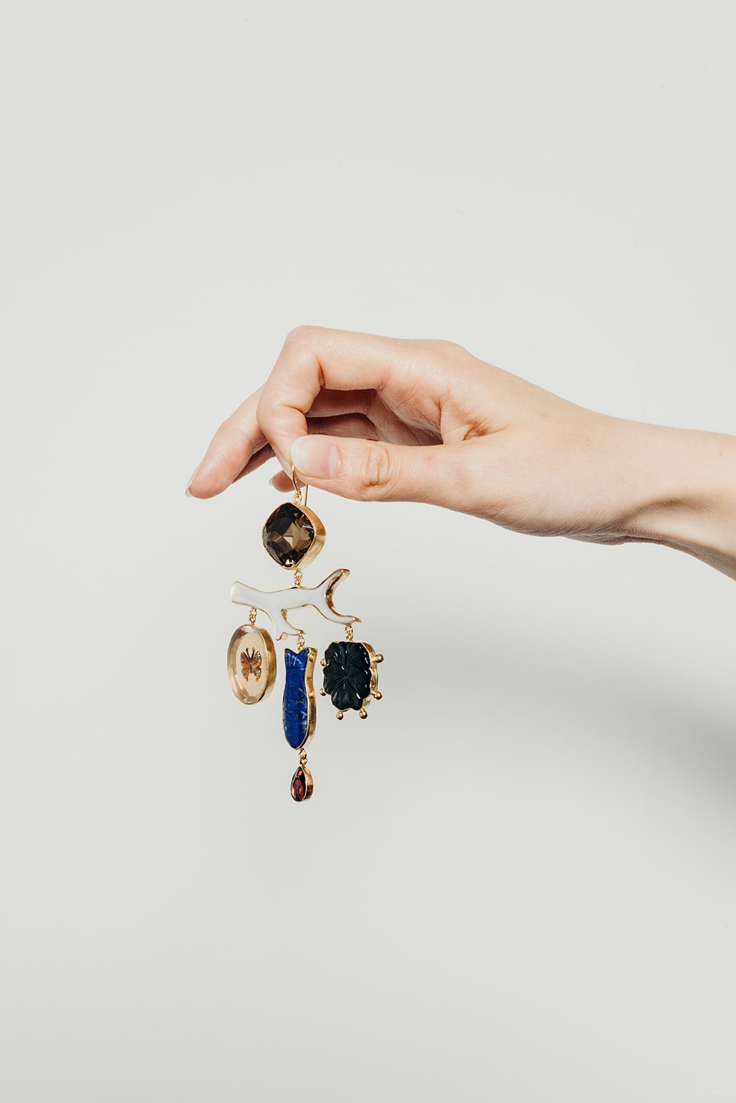 Brand photographer image. Hand holding a gold-toned earring with various gemstones and charms, including a black stone, blue stone, and butterfly charm, against a plain white background.