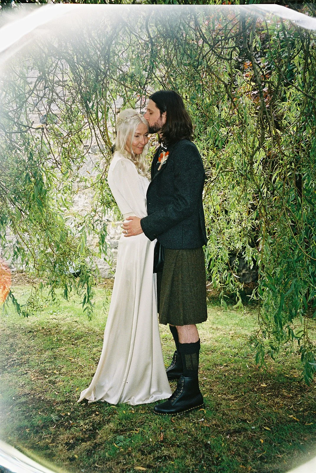 A couple standing under a leafy tree, embracing, with the man kissing the woman's forehead, outdoors during daytime.