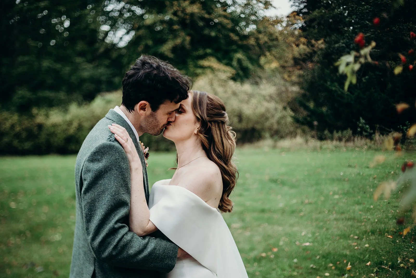A newlywed couple kissing outdoors in a garden, surrounded by green trees and grass.
