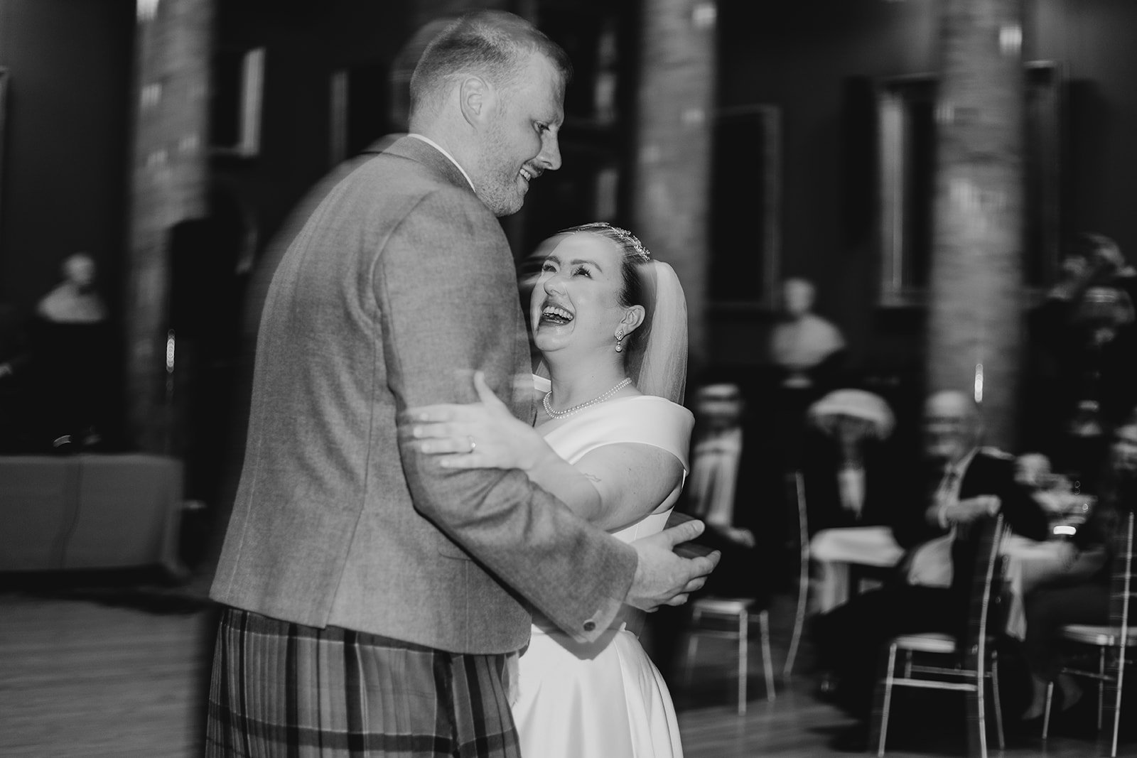 A bride and groom dancing and smiling at their wedding reception, with guests seated in the background.